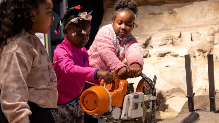 Three girls playing with an interactive, pretending to use power tools to cut into rock.