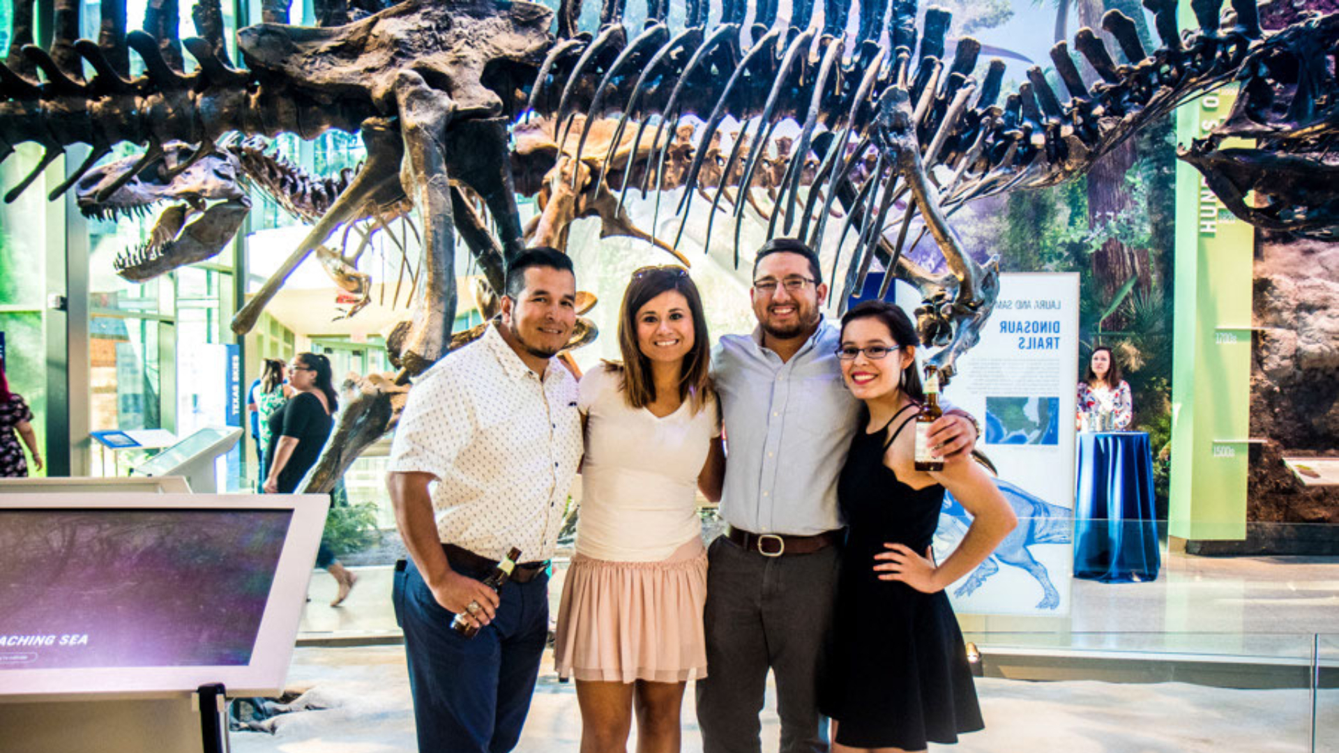 two men and two women standing together and smiling in front of the t.rex. Men are wearing button down short sleeved shirts. Women are wearing tank tops and skirts.