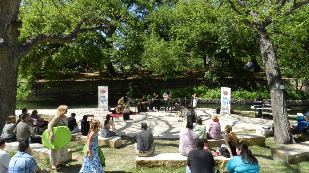 band playing in center of amphitheater, surrounded by crowd sitting on stone benches, with green trees and the river behind them.