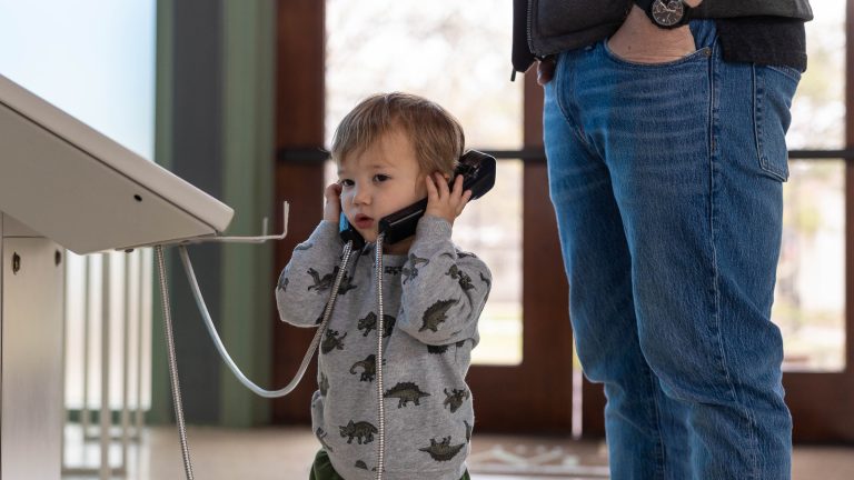 Toddler holds two phones up to his ears inside the dinosaur gallery. He is wearing a shirt with dinosaurs on it.