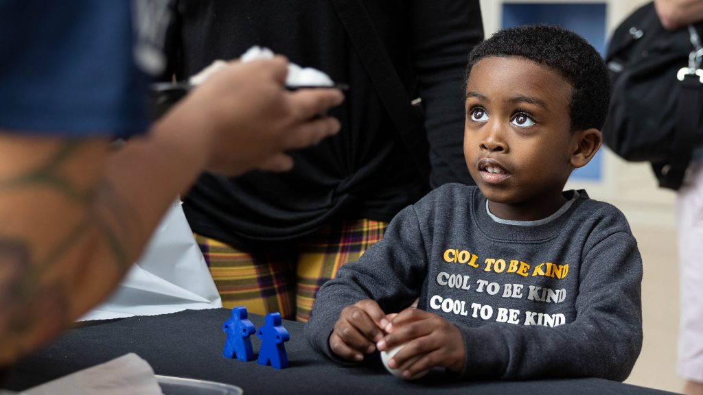 Elementary-aged child holding a ball and looking up at a demonstrator with wide, wondering eyes.
